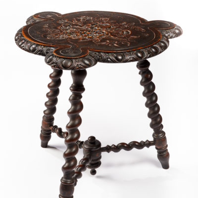Wooden three legged table. Table top is a curved design with three larger and three smaller semi circles. It is decorated in a carved pattern on the surface. The legs are a curved swirling design.