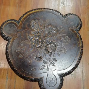 Surface view of a carved wooden table. Flowers, butterflies and dragonflies are carved into it. It is circular in shape with three smaller semi circle protruding from opposing sides.