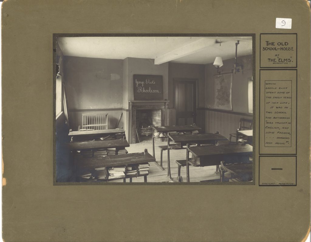 Black and white photograph of an old school room with tables and books underneath. The chalk board at the front has the words George Eliot's School room written on it. The title the old school room, the elms, Nuneaton is written on the right hand side of the green/brown mount.
