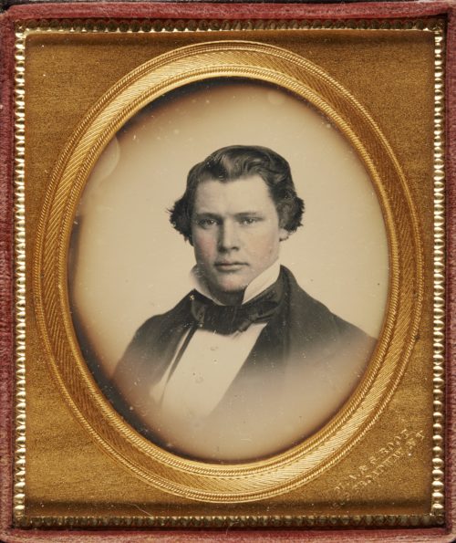 Black and white photograph of a man. The cheeks have been tinted pink. He has a high white collar, white shirt, black necktie and black jacket. The photograph is in an oval frame with a gold mount. It is in a red case.