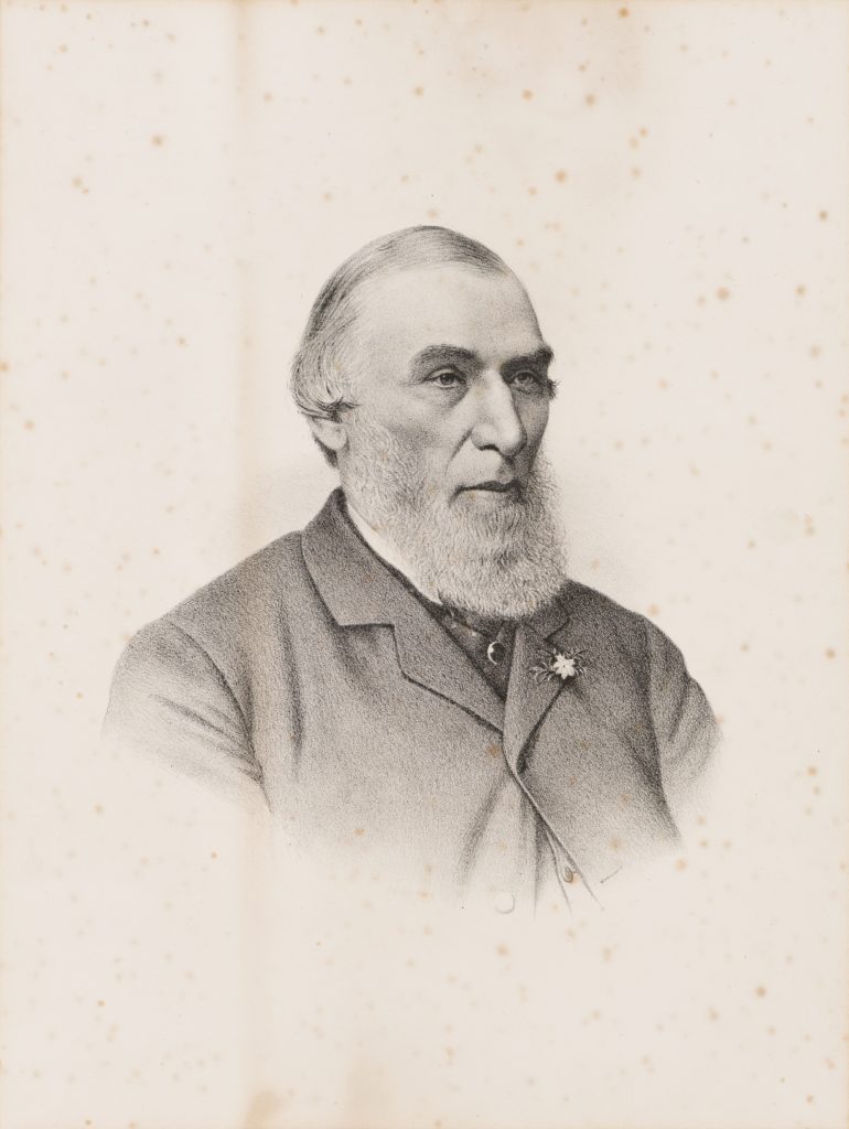 Black and white portrait of a man. He has white hair and a beard. He is wearing a dark suit with a small flower in the capel. The jacket is fastened at the top button. Subject is wearing a necktie and a white collar shirt.