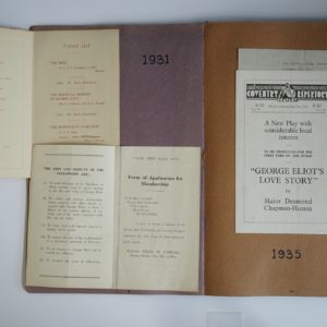 Open scrapbook. On the right hand side a programme for a play, George Eliot's Love Story is stuck onto the page. The date 1935 is written on the page. On the left hand side a menu and toast list and membership application form are stuck onto the page. The date 1931 is written on the page.
