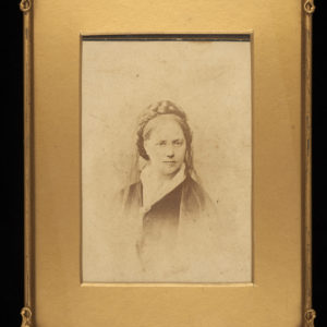 A black and white photograph of Rufa Hennell. It shows a woman whose hair is plaited around the top. There is a light veil around the back of her head. She is wearing a white collar and dark cloak or shawl fastened with a broach. The photograph as a gold border and frame.