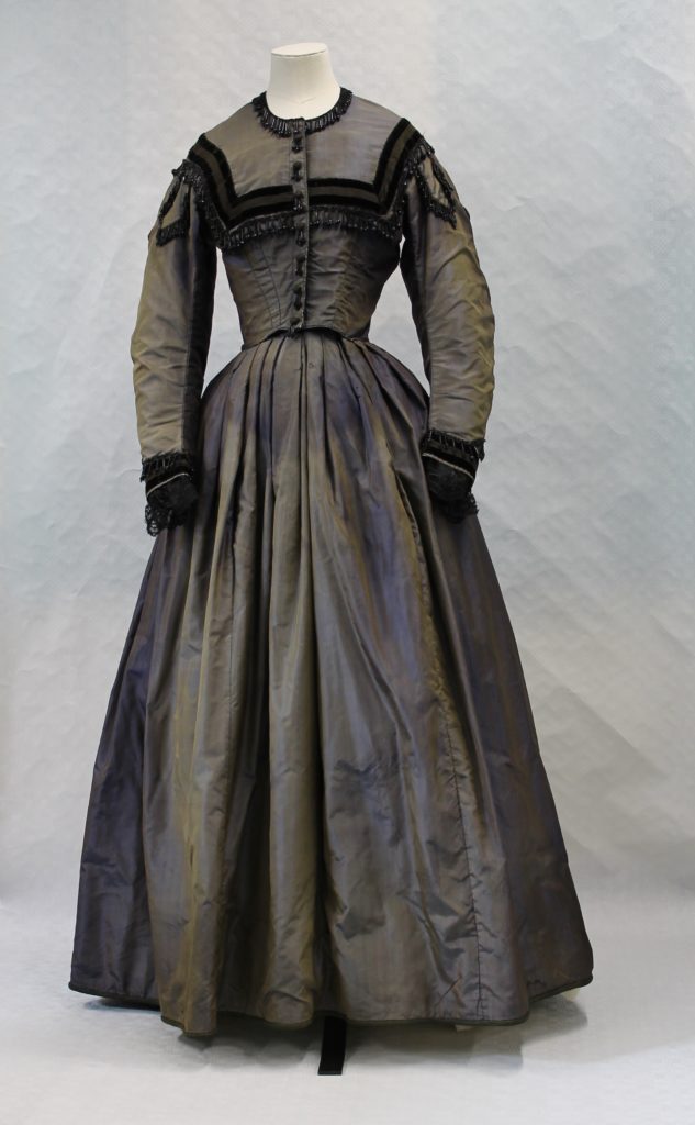 Mannequin displaying a blue floor length silk dress with long sleeves. The bodice has a square shaped black velvet trim. There is black bead trim round the neck following a black velvet trim. There is black velvet trim and lace at the wrists. There are buttons along the front to the waist.