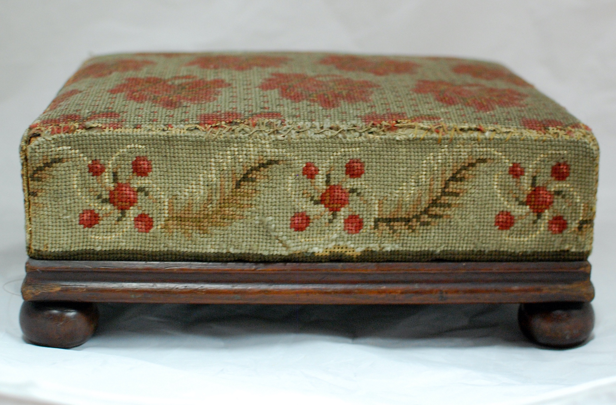 Side view of a wooden footstool with embroidered cushion. It is embroidered with a red flower pattern.