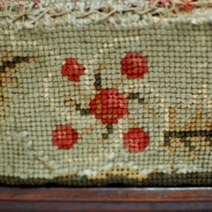Detail of a red pattern on the side of an embroidered footstool. Pattern consists of four red dots with a larger red dot in the centre with swirls coming from it.