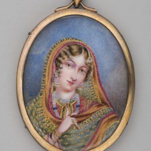 An oval shape pendant with a gold frame around a painting of a woman. She is wearing a highly patterned shawl which she holds with one hand. She is wearing earrings, necklaces and bracelets.