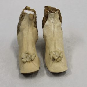 Front view of two small cream coloured kid leather boots with silk cream bows near to the toe. They have a small heel. The sides have a darker band of smocking to allow easier fitting.