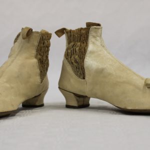 Two small cream coloured kid leather boots with silk cream bows near to the toe. They have a small heel. The sides have a darker band of smocking to allow easier fitting.