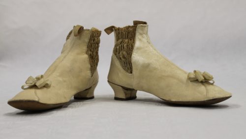 Two small cream coloured kid leather boots with silk cream bows near to the toe. They have a small heel. The sides have a darker band of smocking to allow easier fitting.
