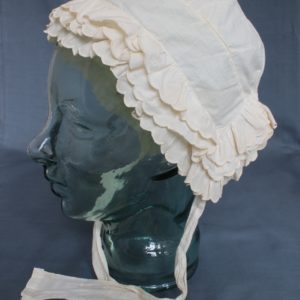 Side view of a white bonnet displayed on a clear mannequin. There are three rows of scalloped edged bands around the front and back. It has two lengths of cotton which would tie under the chin.