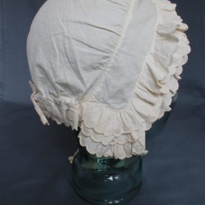 Right side view of a white bonnet displayed on a clear mannequin. There are three rows of scalloped edged bands around the front and back. It has two lengths of cotton which would tie under the chin.