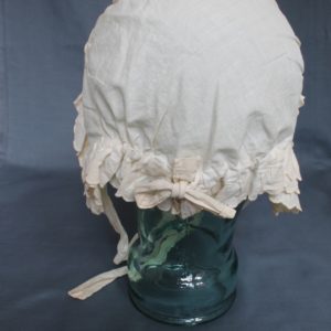 Back view of a white bonnet displayed on a clear mannequin. There are three rows of scalloped edged bands around the front and back. It has two lengths of cotton which would tie under the chin.