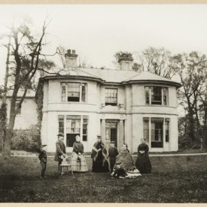 Black and white photograph showing a large white house with five windows and a doorway. There are trees to the left and right of the house. There is a large lawn in front of the house and a group of people gathered on it in front of the house.