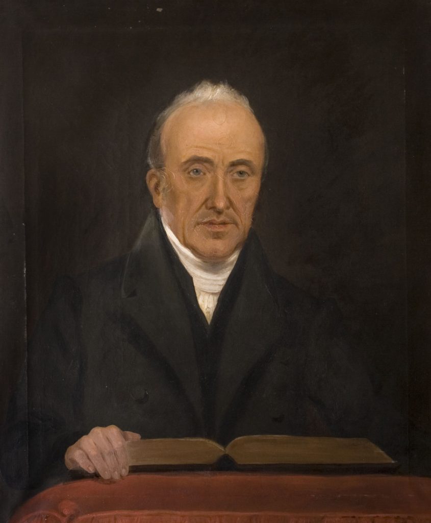 Painting of a man with white grey hair. He is wearing a white clerical collar and black robe. He is standing with one hand on a open book which is reading on a pedestal.