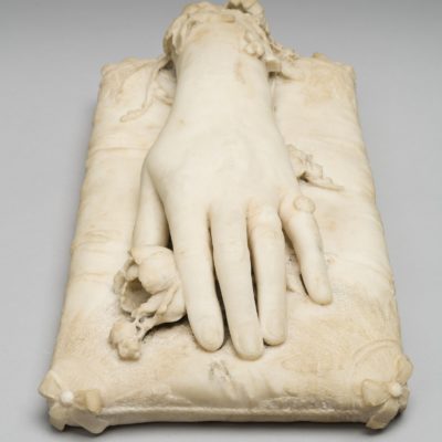 Front view of a white marble sculpure of a hand resting on a marble cushion. Under the hand a marble rose is visible.
