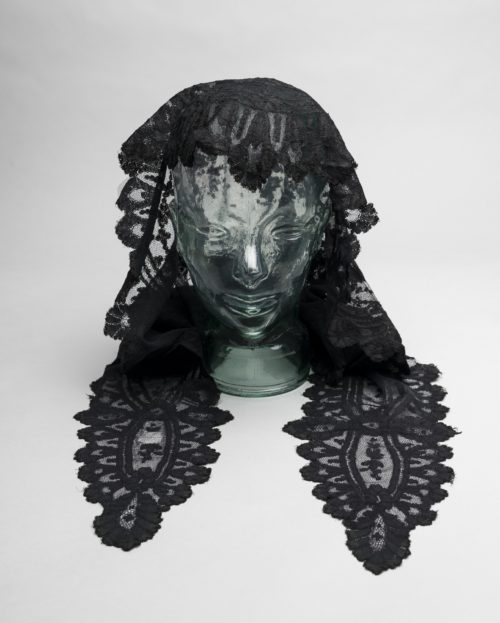 Black lace head shawl displayed on a glass mannequin. It covers the back of the head and has two long pieces on each side.