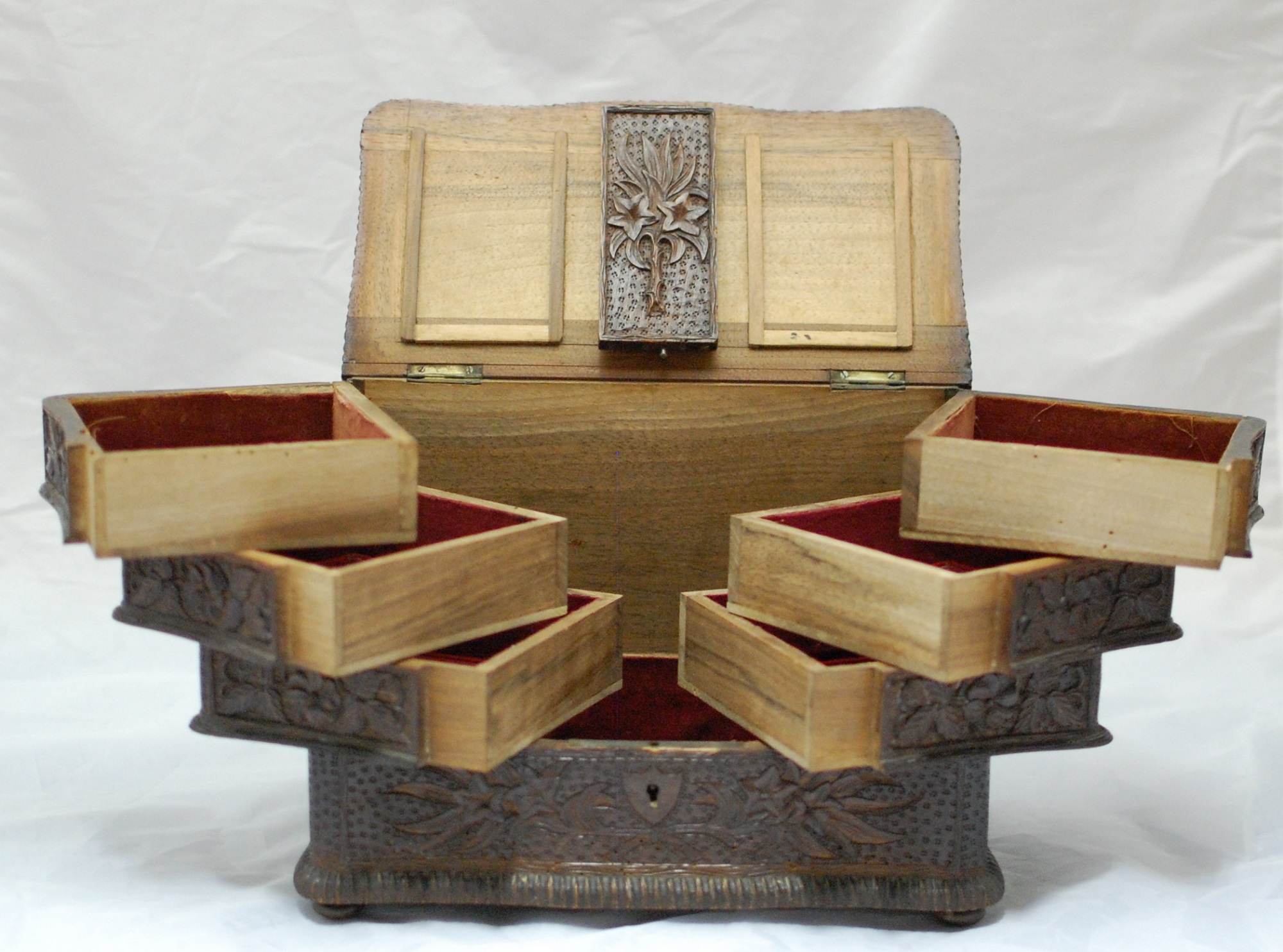 Open view of a wooden lace box. The open lid is of page wood with a dark wood decorated panel in the centre. There are three stacked compartments on the left and right which are lined with a red material.. The centre provides a large storing space.