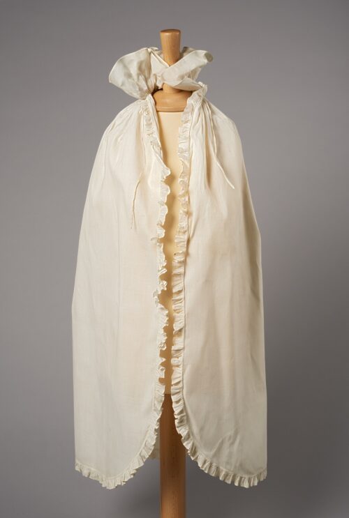 Photograph of a cream cloak with collar with ties at the collar. Displayed on a mannequin.